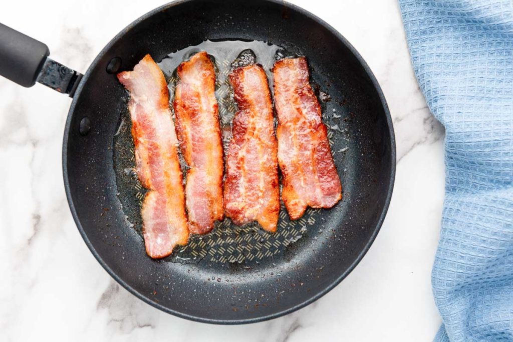 Bacon sizzling in a frying pan on a marble countertop.