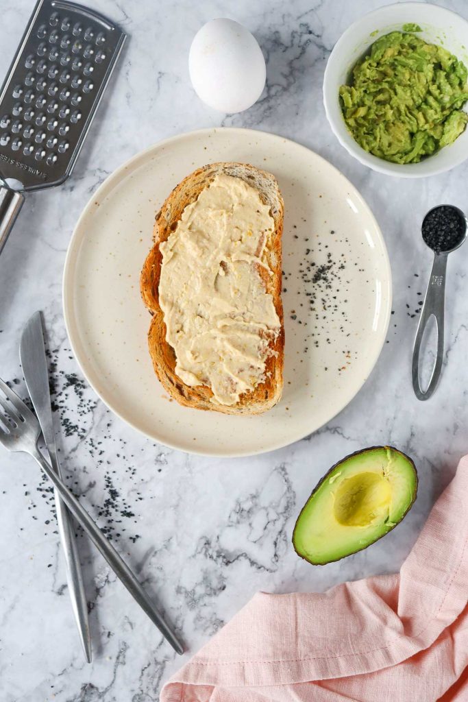 Avocado toast with hummus on a white plate with a knife and utensils.