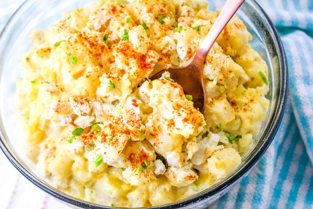 A glass bowl of egg potato salad topped with chopped chives and paprika, with a fork resting inside the mixture.