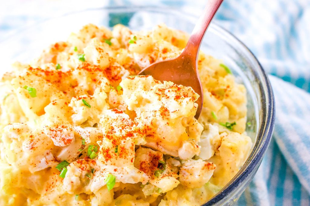 A bowl of creamy potato salad garnished with paprika and green herbs, with a spoon inside.