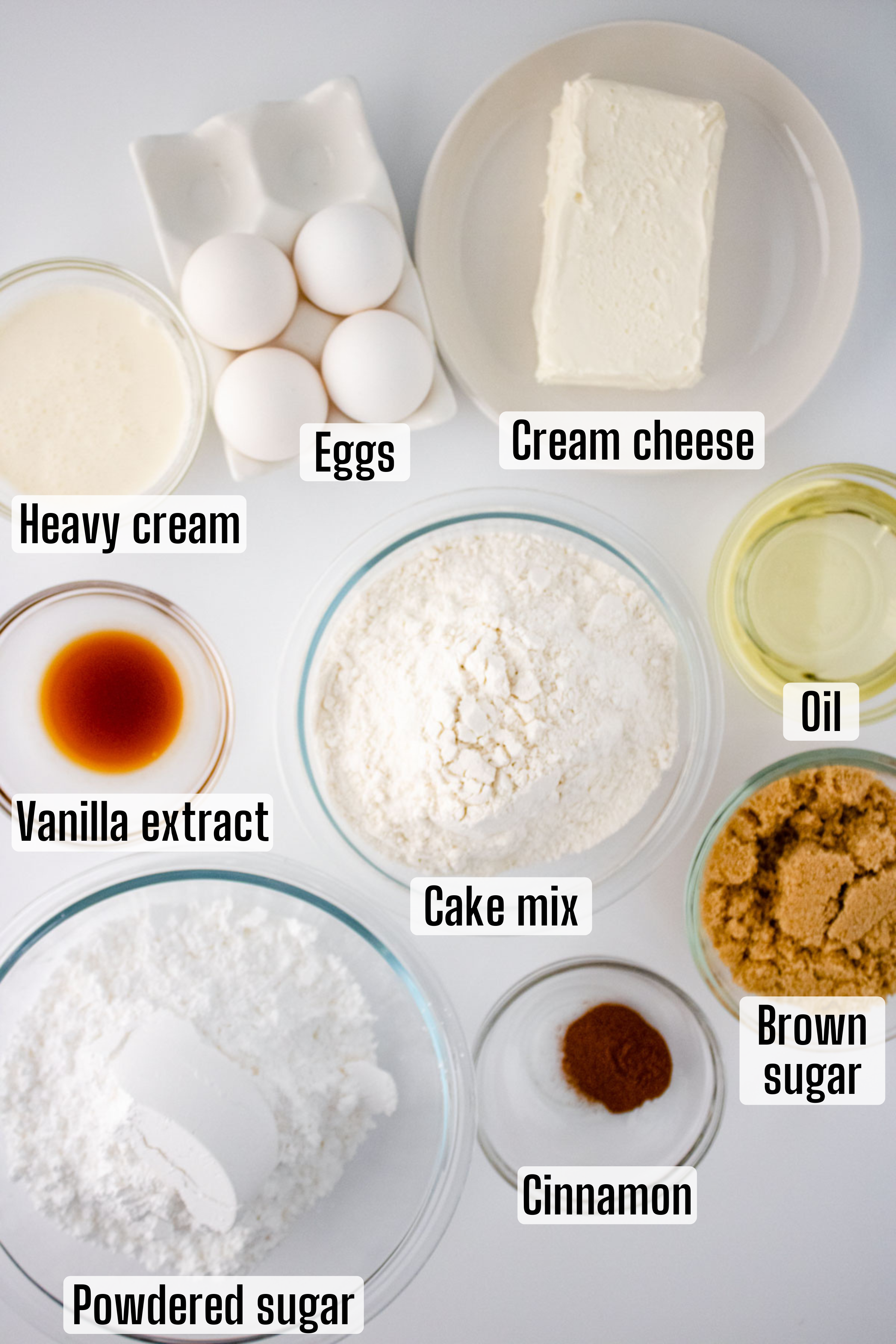 Various labeled ingredients for a honey bun cake recipe, including eggs, cream cheese, heavy cream, oils, and spices, are arranged neatly on a white surface.