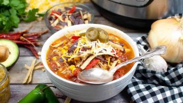 A bowl of tortilla soup topped with cheese and jalapeños, surrounded by ingredients and a slow cooker in the background.