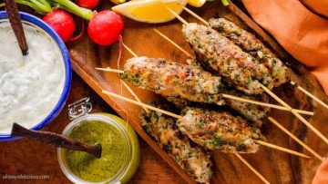 Grilled chicken skewers on a wooden board, served with radishes, lemon, and dipping sauces.