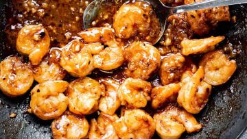 Shrimps coated in a dark, glossy sauce, garnished with herbs, served in a skillet with a spoon.