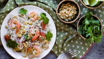 A plate of shrimp and glass noodle salad garnished with cilantro, lime, and peanuts, on a patterned tablecloth.