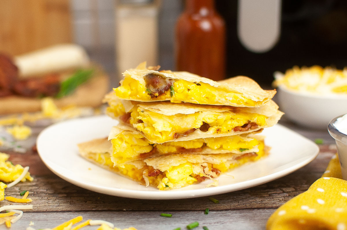 A stack of breakfast quesadillas filled with eggs and bacon on a white plate, with hot sauce and a bowl of cheese in the background, perfect for exploring new breakfast recipes.