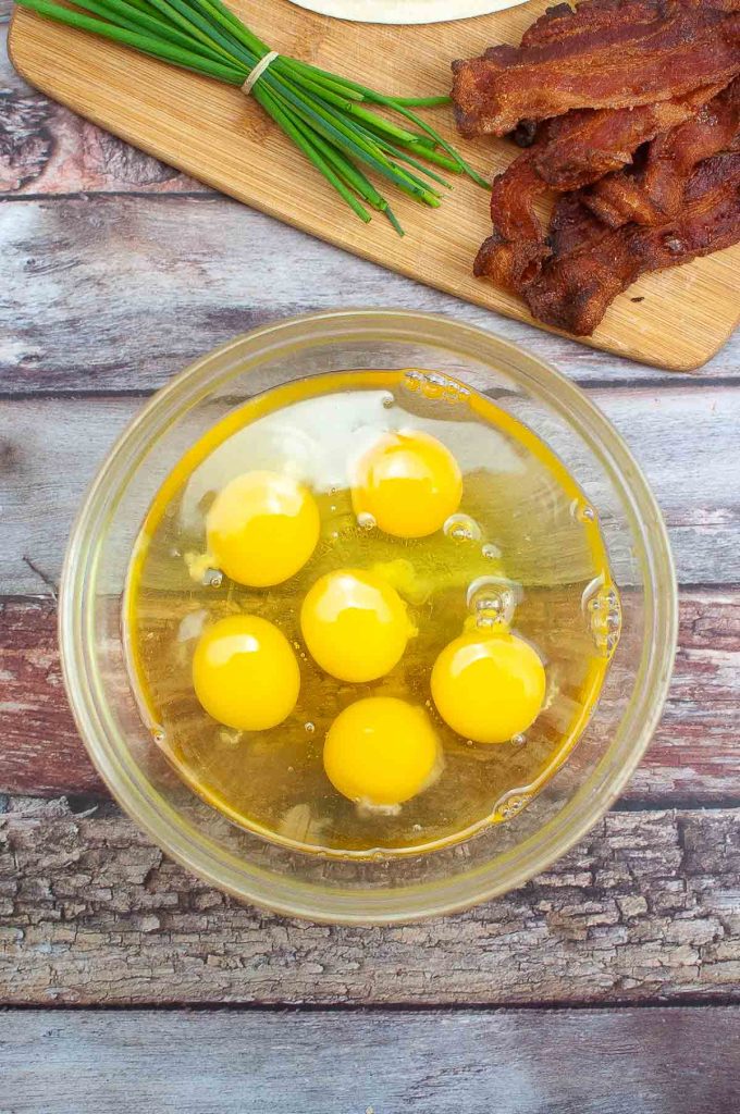 A glass bowl containing six raw eggs with yolks intact, set on a wooden surface with a bunch of chives and cooked bacon strips in the background, prepared for making Breakfast Quesadillas.