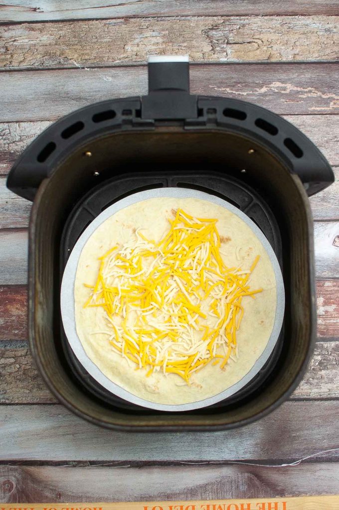 A tortilla with shredded cheese on top inside an open air fryer, ready to become breakfast quesadillas.