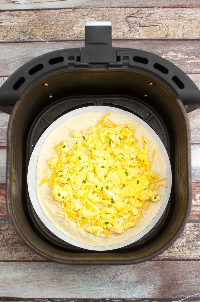 A Breakfast Quesadilla in an air fryer basket topped with shredded cheese and scrambled eggs ready to be cooked.
