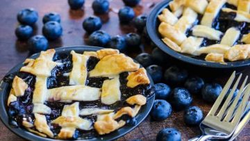 Two mini blueberry pies with lattice crusts on black plates surrounded by fresh blueberries, with a fork on the side.