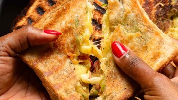 Close-up of hands with red nails holding a grilled cheese sandwich cut in half, showing melted cheese and fillings.