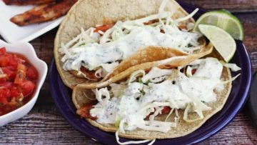 Three fish tacos with cabbage slaw and creamy sauce on a purple plate, served with lime wedges and salsa on the side.