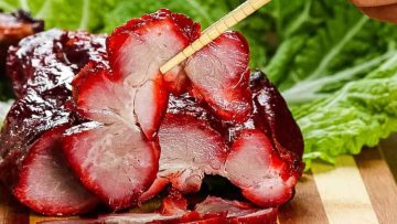 Sliced char siu pork with a shiny glaze, being picked up with chopsticks, placed next to green lettuce leaves on a wooden board.