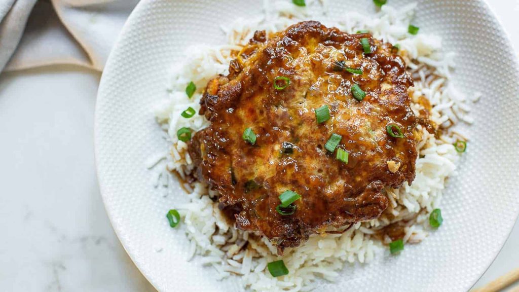 A plate of white rice topped chicken egg foo young, garnished with chopped green onions.