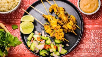 Grilled Thai chicken satay skewers served with a side of cucumber salad and lime slices on a black plate, with rice and dipping sauce in the background.