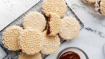 Dulce de Leche Cookies with cream filling on a wire rack, one broken in half to show caramel inside, next to a jar of caramel.