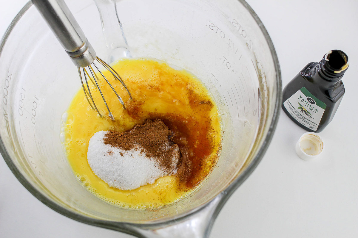 Mixing ingredients for a French Toast Casserole in a glass bowl with a whisk, alongside a bottle of vanilla extract.