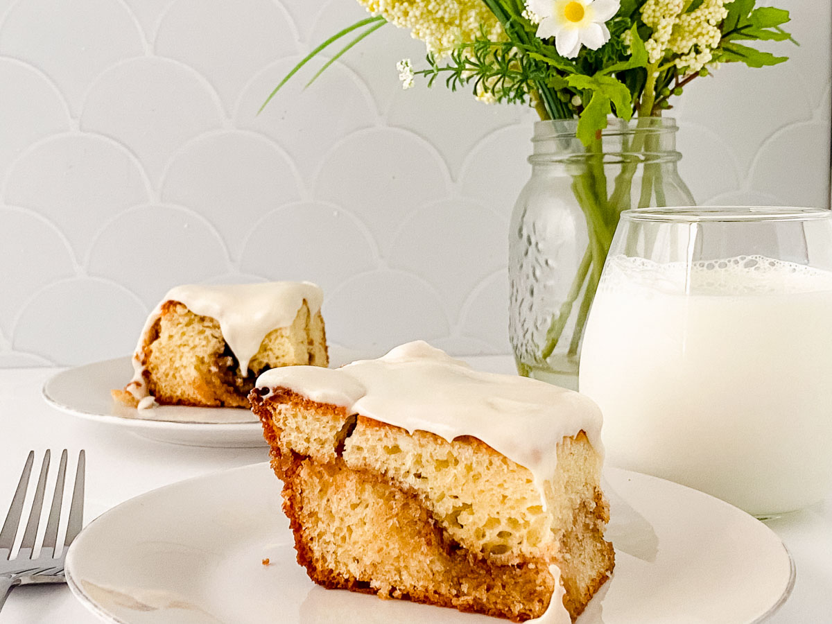 Two slices of honey bun cake on white plates with icing, accompanied by a glass of milk, set against a tiled backsplash.