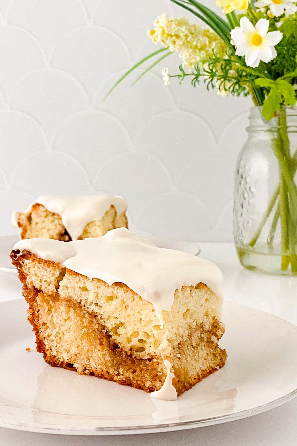 A slice of honey bun cake with creamy icing on a white plate, background featuring a jar of yellow and white flowers.