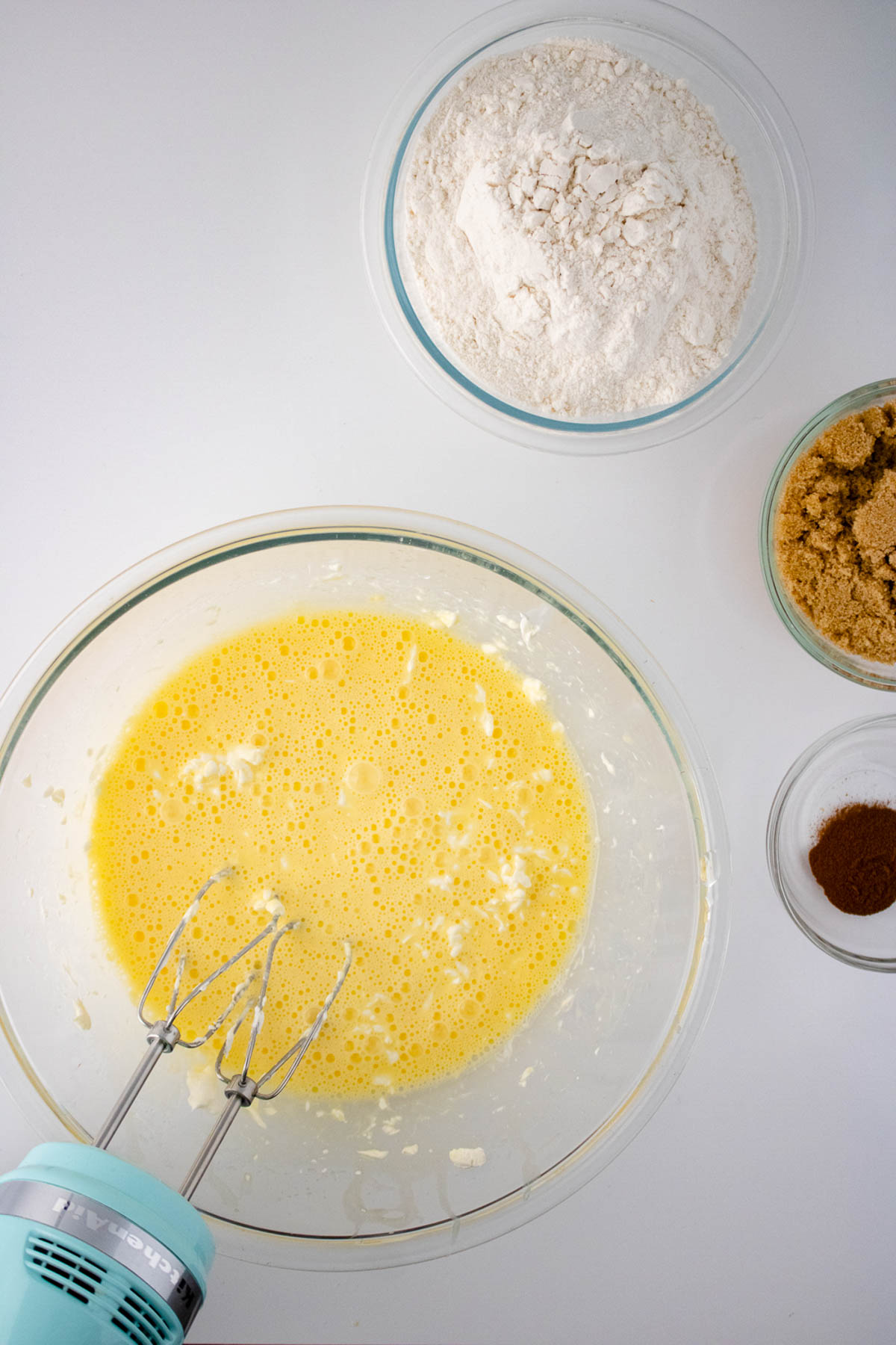 Top view of a kitchen counter with ingredients for dessert: a bowl of whisked eggs, bowls of flour, brown sugar, and cocoa powder, and a hand mixer.