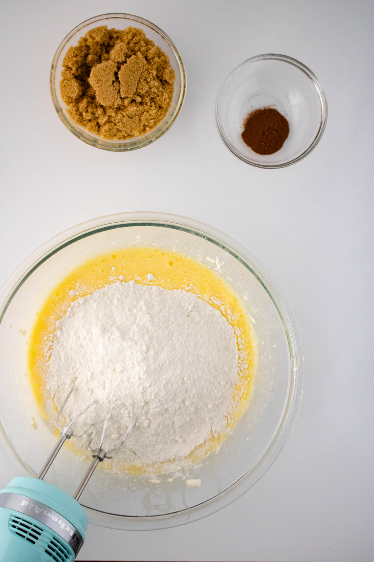 Overhead view of ingredients for a honey bun cake, featuring a bowl with flour and egg mixture, accompanied by bowls of brown sugar and cinnamon, with a hand mixer in use.