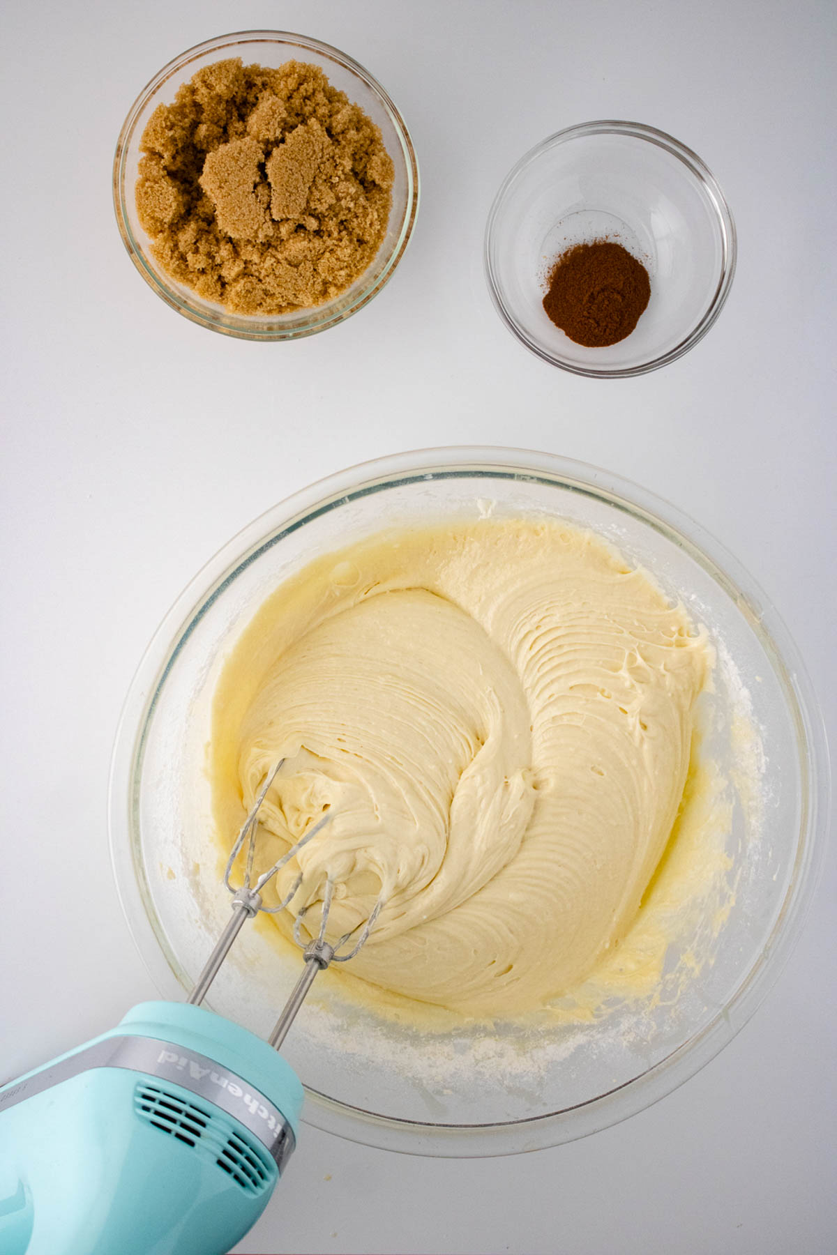 Top view of a bowl containing creamed batter for honey bun cake with a hand mixer, flanked by bowls of brown sugar and cocoa powder on a white surface.