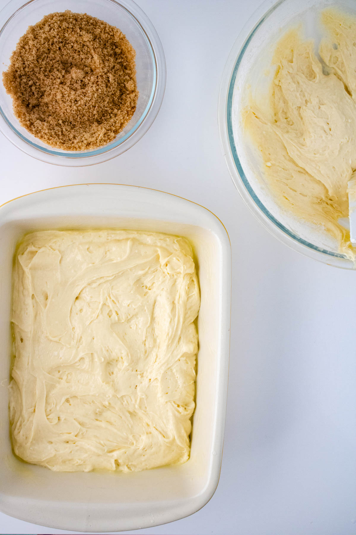 Top view of a baking dish with uncooked honey bun cake batter, a bowl with creamy mixture, and a bowl of brown sugar, all on a white surface.
