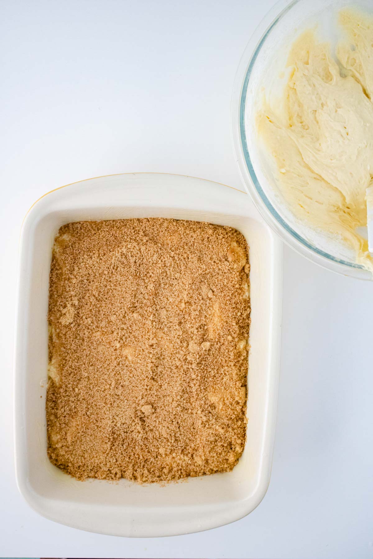 A square baking dish filled with a crumbly brown sugar mixture beside a glass bowl containing honey bun cake batter.