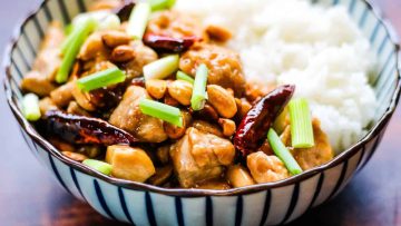 A bowl of kung pao chicken with peanuts, chili peppers, and green onions, served alongside white rice.