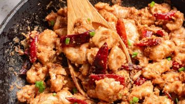 Stir-fried peanut butter chicken with red chili peppers and green onions in a black skillet, being mixed with a wooden spatula.