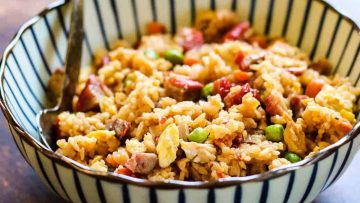 A bowl of pork fried rice with peas, chunks of sausage, and scrambled eggs, served with a spoon, on a wooden table.