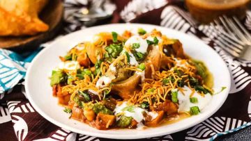A plate of samosa chaat garnished with yogurt, green chilies, cilantro, and sev, served on a patterned tablecloth.