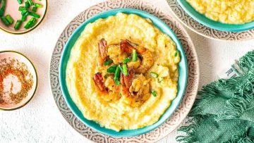 A plate of creamy grits topped with sautéed shrimp and garnished with green onions, served on a decorative plate with a green napkin and small bowl of spices on the side.