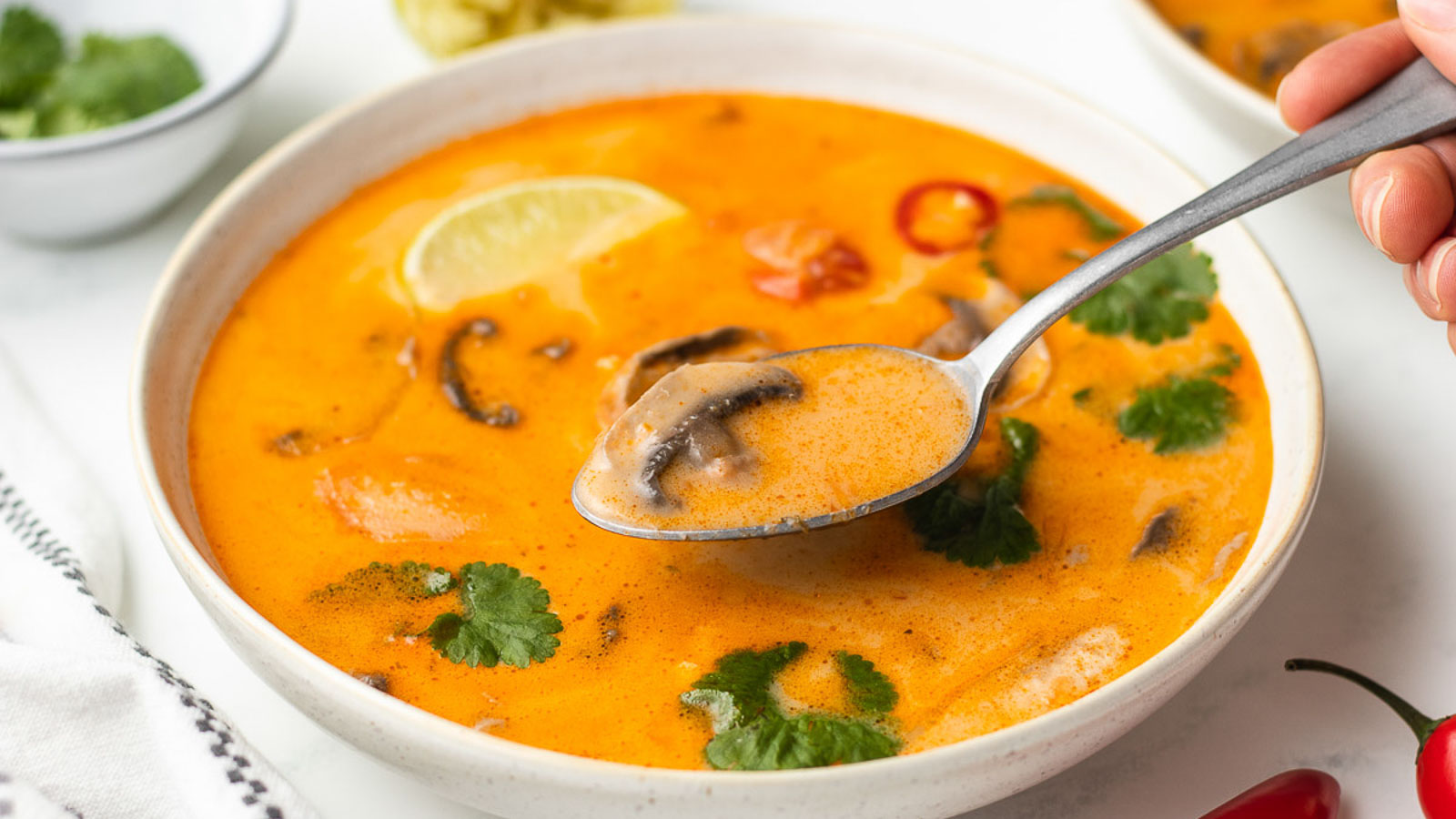 A bowl of soup garnished with cilantro, lime, and chili slices, with a person taking a spoonful.