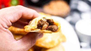 A close-up image of a hand holding an air fried empanada with visible meat filling.