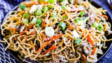 A plate of stir-fried noodles with beef, carrots, and green onions, garnished with sesame seeds.