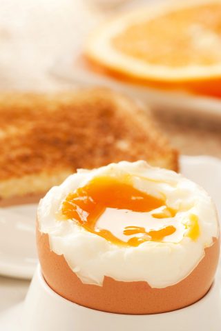 A soft-boiled egg in an egg cup with a runny yolk, made using an air fryer, accompanied by a slice of buttered toast and half an orange in the background.