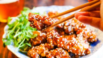 Chopsticks holding a piece of sesame-coated fried chicken over a plate of similar pieces, with greens on the side and a glass of beer in the background.