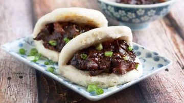 Two Chinese pork belly buns served on a patterned plate, garnished with green onions, with a side of sauce in the background.
