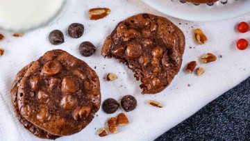 Chocolate cookies with nuts and candies on a white napkin, one cookie broken in half, with a glass of milk in the background.