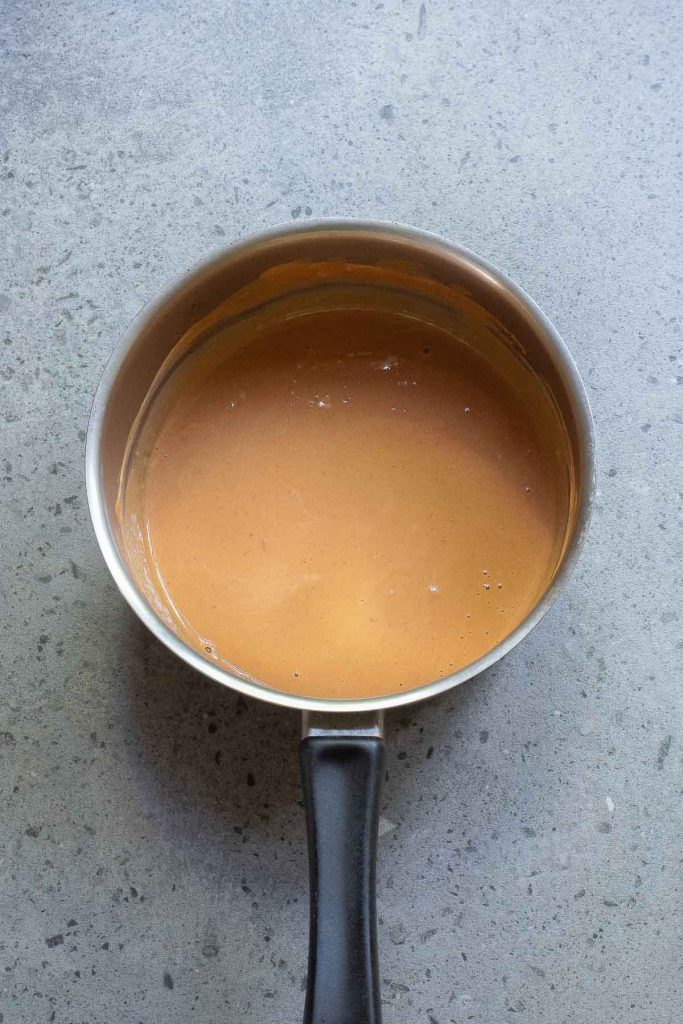 A saucepan with light brown peanut sauce and savory bits of bacon against a speckled gray background.
