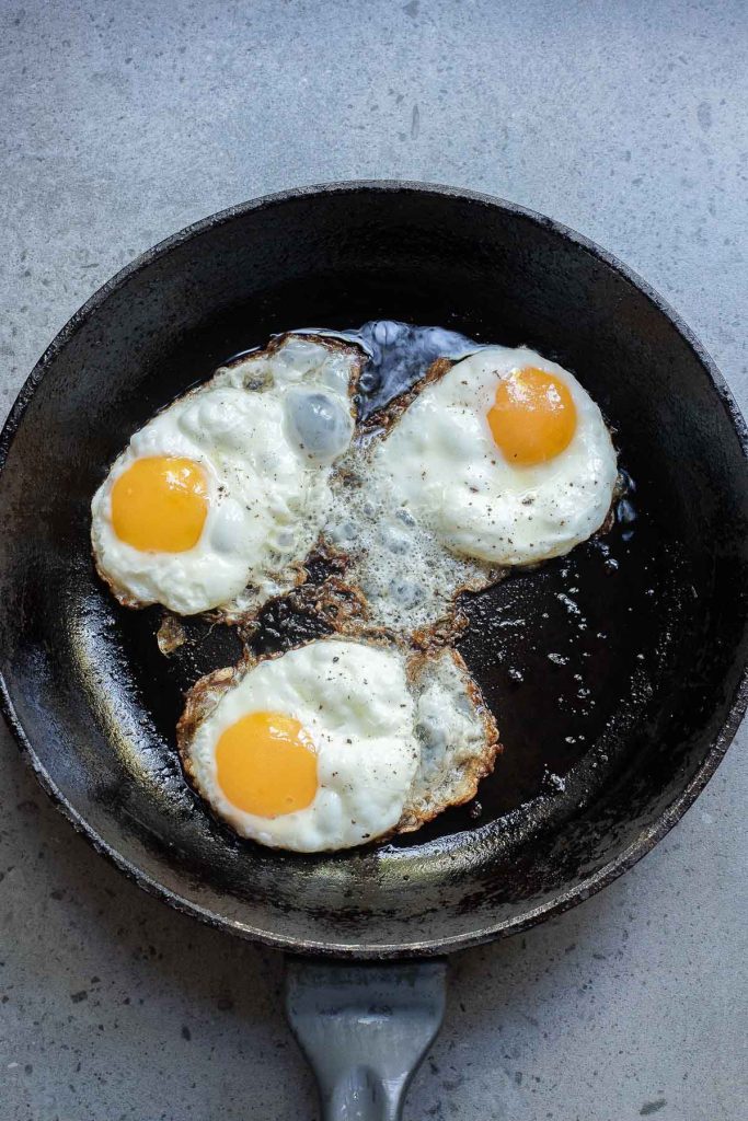 Three sunny-side-up eggs sizzling in a cast iron skillet on a gray surface, with crispy bacon adding an irresistible aroma.
