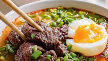 A bowl of ramen with beef, a soft-boiled egg, green onions, and chopsticks.