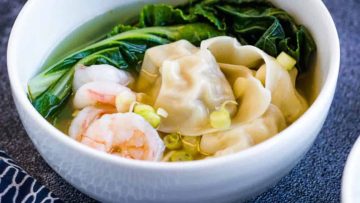 A bowl of Asian dumpling soup with shrimp, greens, and scallions, presented on a textured blue tablecloth.