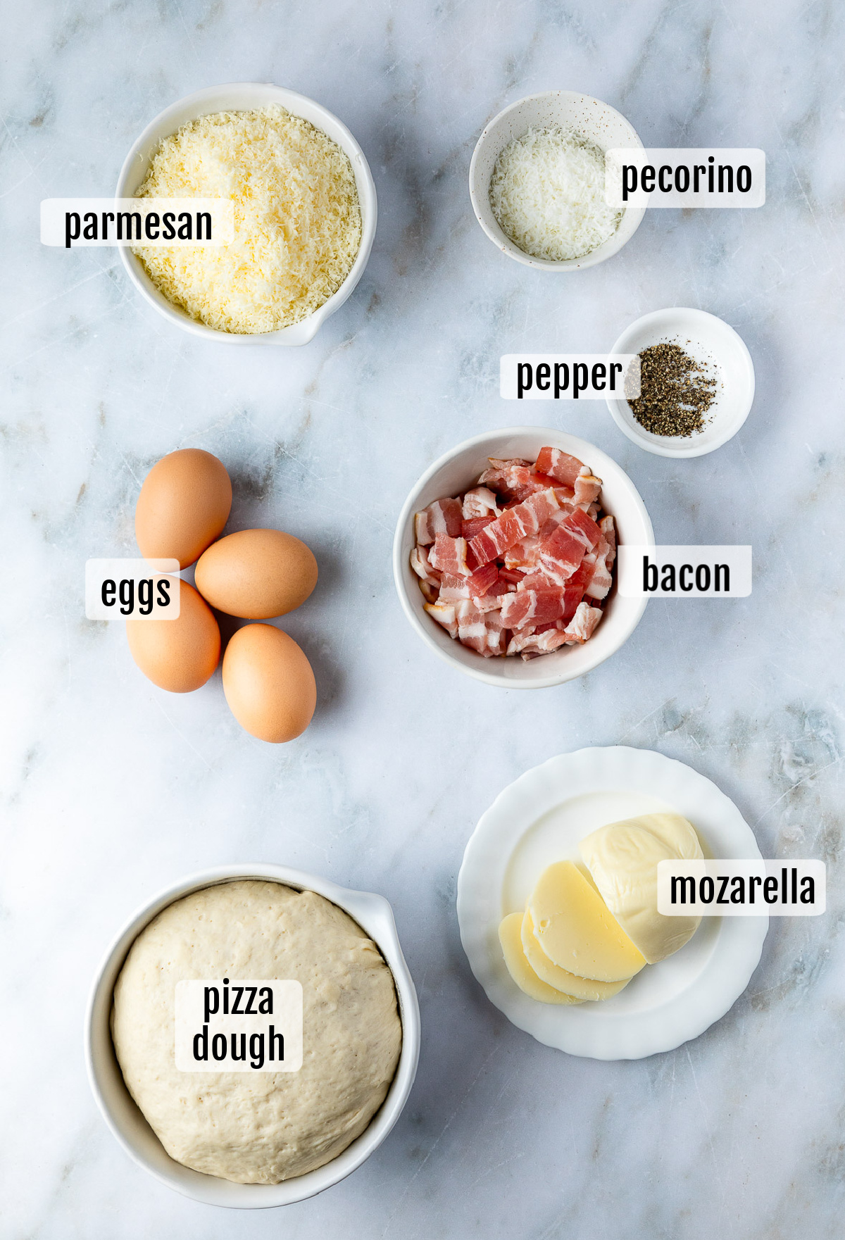 An arrangement of cooking ingredients for Pizza Carbonara includes Parmesan, pecorino, pepper, eggs, bacon, mozzarella, and pizza dough, each labeled accordingly on a marble surface.
