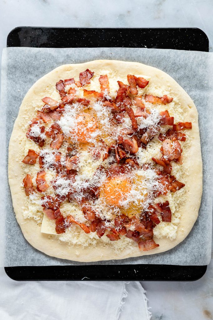 A delightful carbonara pizza creation awaits: unbaked and topped with crispy bacon, shredded cheese, and two raw eggs, all resting on parchment paper.