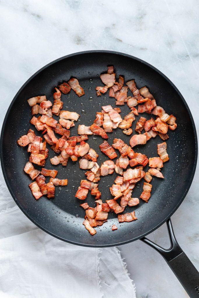 Diced bacon pieces sizzle in a black nonstick pan on a marble surface, infusing the air with a tantalizing hint of carbonara pizza.