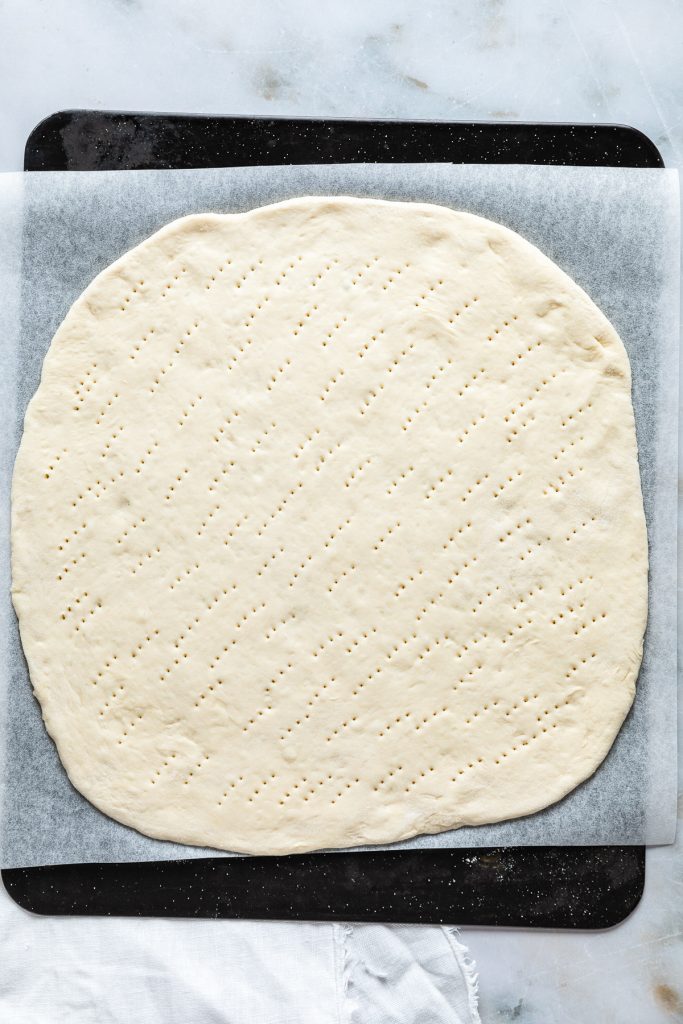 A rolled-out carbonara pizza dough on parchment paper, with a perforated surface, sits elegantly on a baking tray.