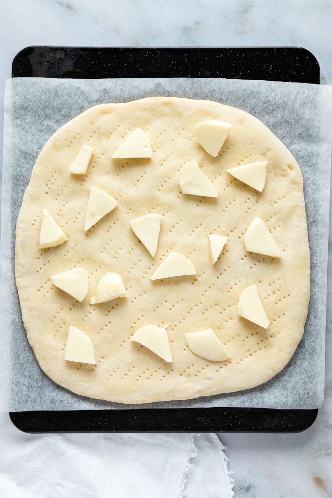 Carbonara pizza dough topped with scattered cheese pieces rests on a baking tray lined with parchment paper.
