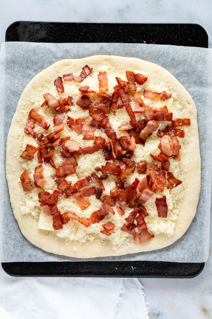A delightful carbonara pizza creation: a square dough topped with shredded cheese and bacon pieces, all set on parchment paper, ready for baking.